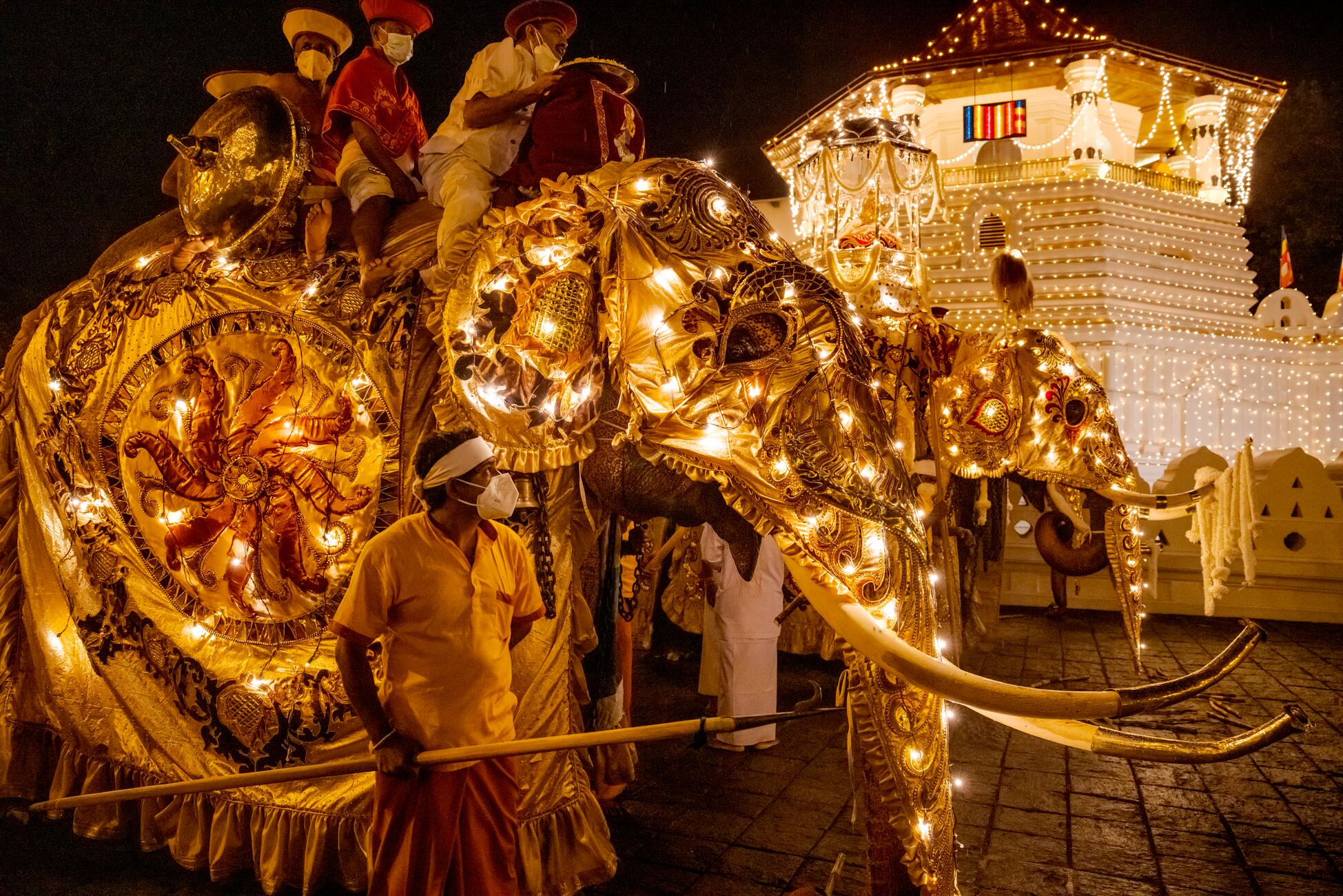Picture of two elephants at night wearing elaborate costumes with lights being ridden by people